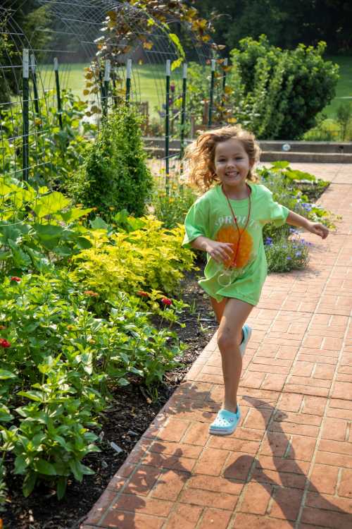 A young girl with curly hair runs joyfully along a garden path, surrounded by vibrant flowers and greenery.