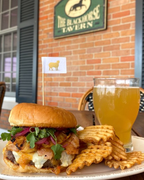 A gourmet burger topped with grilled pineapple, cilantro, and pickled onions, served with waffle fries and a drink.