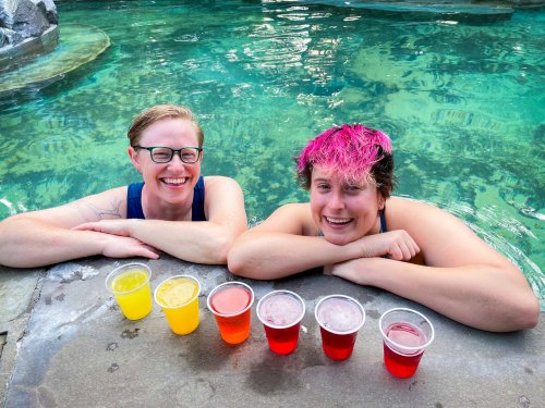 Two smiling women in swimsuits relax by a pool, with colorful drinks in front of them on the edge.