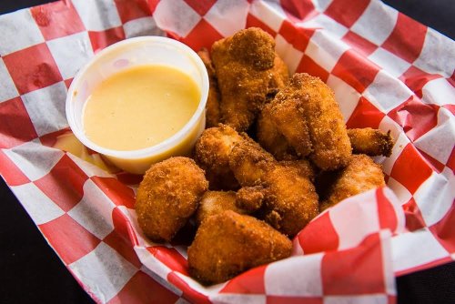 A basket of golden-brown fried chicken pieces with a small cup of dipping sauce, lined with red and white checkered paper.