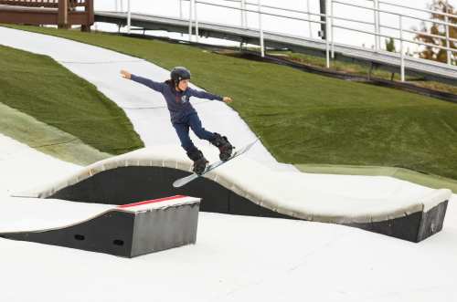 A child in a helmet performs a trick on a snowboard at a snowboarding park with artificial slopes.
