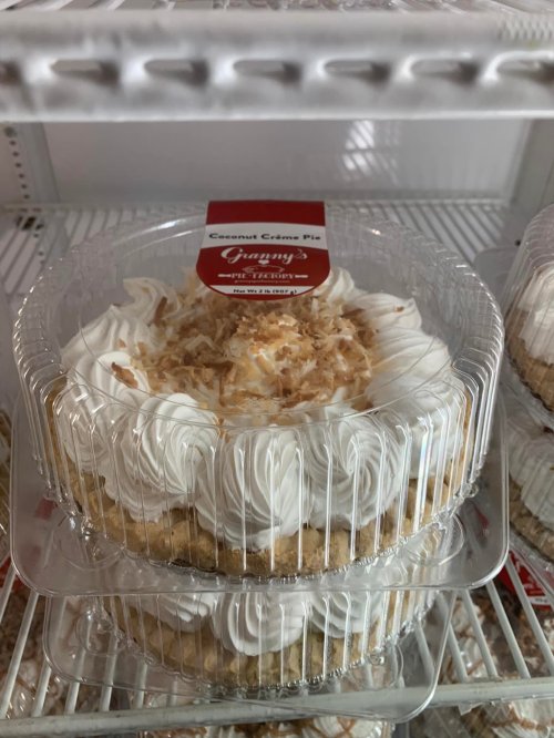 Two coconut cream pies in clear plastic containers, topped with whipped cream and toasted coconut flakes.