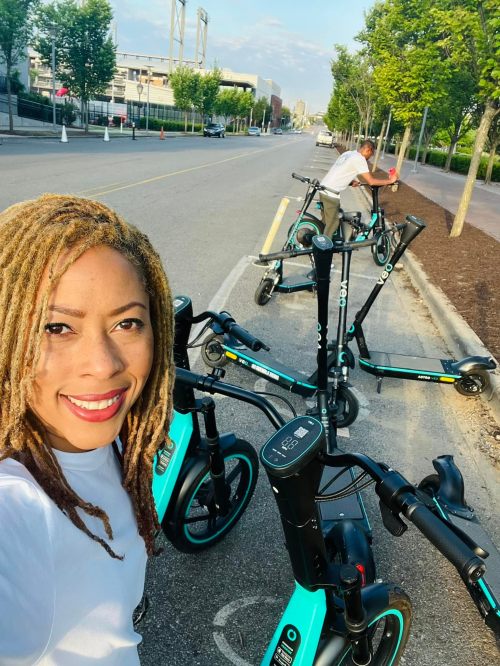 A woman with long, curly hair smiles in front of electric scooters parked on a city street. A man adjusts a scooter nearby.