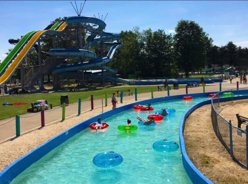 A water park scene with colorful inner tubes floating in a lazy river, surrounded by slides and green trees.