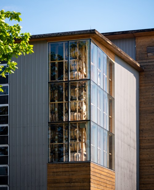 Modern building with large glass windows showcasing stacked barrels inside. Green foliage in the foreground.