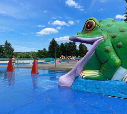 A large, colorful frog sculpture with a long tongue sits beside a water play area under a blue sky.