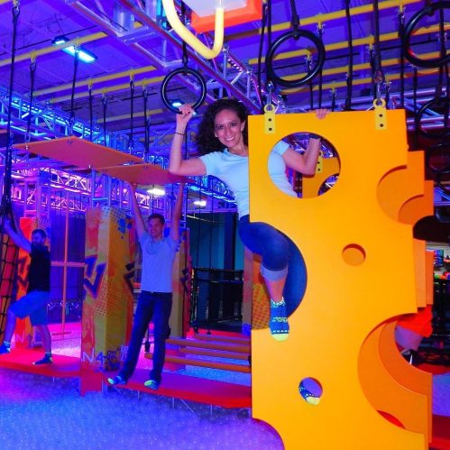A vibrant indoor play area with colorful obstacles; a woman swings from a ring while two others navigate the course.