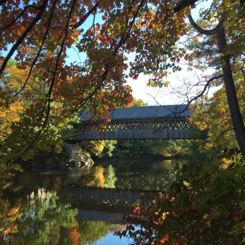 A wooden covered bridge spans a calm river, surrounded by vibrant autumn foliage and reflections in the water.