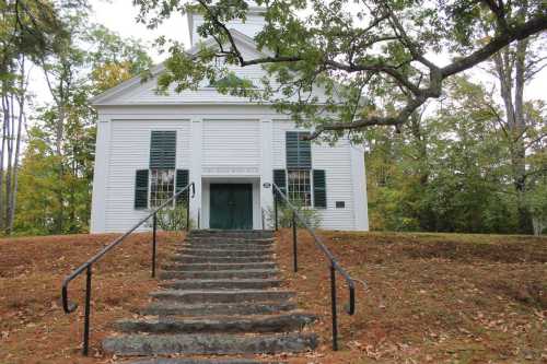 A white building with green doors and shutters, surrounded by trees and a grassy area, with steps leading up to it.