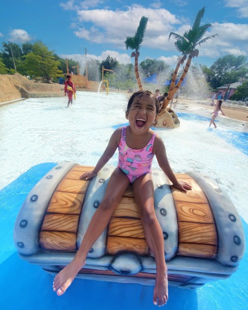 A joyful child in a swimsuit sits on a treasure chest at a water park, surrounded by palm trees and splashing water.