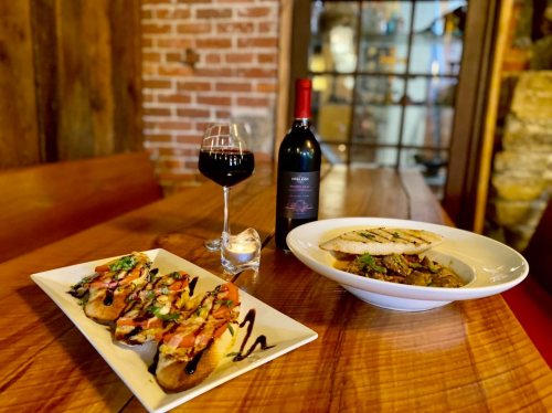A wooden table displays a plate of bruschetta, a bowl of stew, and a glass of red wine in a cozy setting.