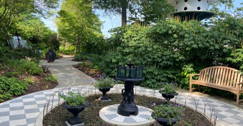 A serene garden path with lush greenery, a decorative fountain, and a wooden bench surrounded by flowers.