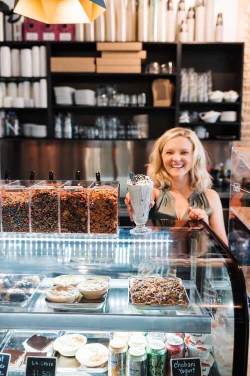 A smiling woman stands behind a glass display, holding a colorful sundae with toppings, surrounded by various snacks.