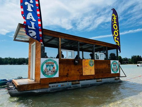 A wooden floating restaurant with flags, docked on a lake under a blue sky.