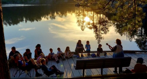 A group of people sitting by a lake at sunset, listening to a musician playing guitar on a wooden dock.