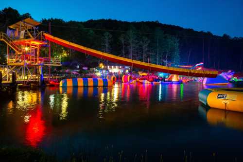 Colorful water park at night with a large slide, inflatable obstacles, and illuminated surroundings reflecting on the water.