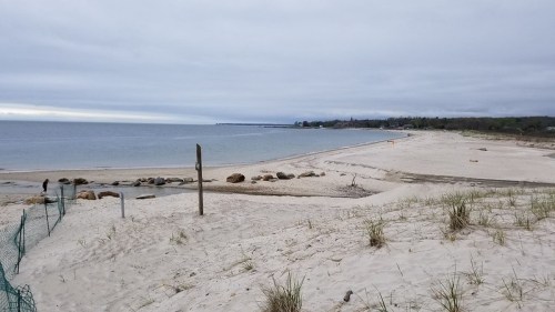 A sandy beach with gentle waves, rocky shoreline, and overcast skies in the distance.