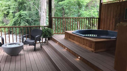 A cozy outdoor deck featuring a hot tub, seating area, and lush greenery in the background.