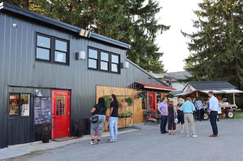 A group of people gathers outside a modern black building with red accents, surrounded by trees and outdoor seating.