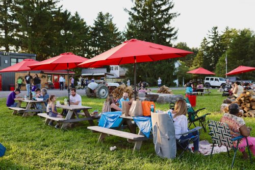 People enjoying a gathering outdoors under red umbrellas, with picnic tables, grass, and trees in the background.