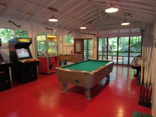 A game room featuring a pool table, arcade machine, foosball table, and a claw machine, with large windows and red flooring.