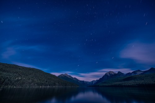 A serene night landscape featuring a calm lake, mountains, and star trails in a deep blue sky.