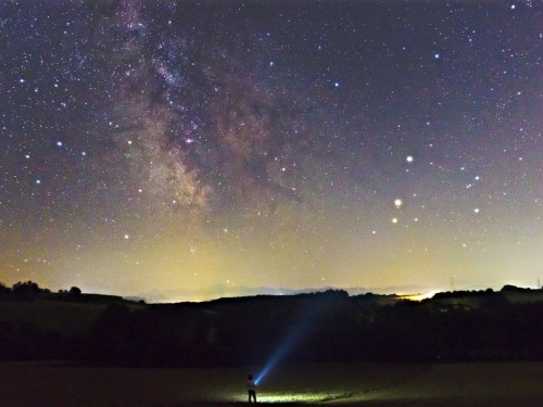 A person stands in a field, shining a flashlight at the starry night sky filled with the Milky Way and distant planets.