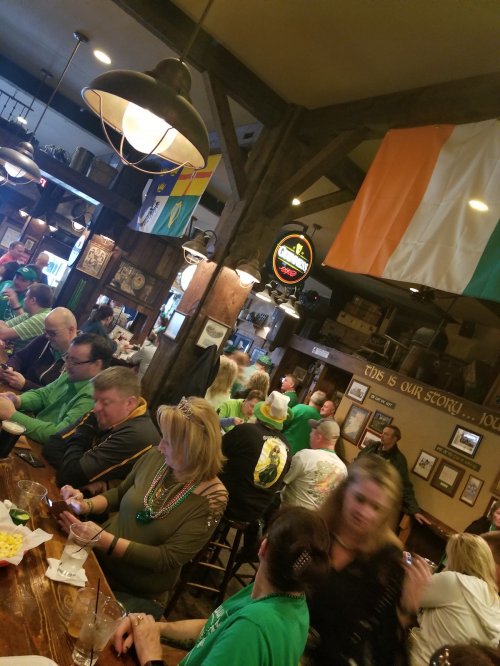 A lively pub scene with patrons in green attire, enjoying drinks and celebrating, decorated with Irish flags.
