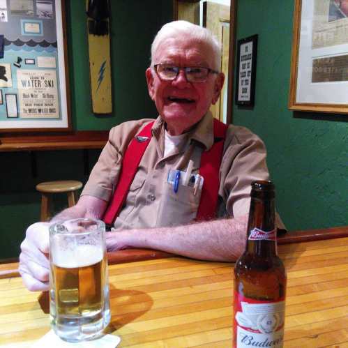 An older man with glasses smiles at the bar, holding a beer, with a bottle of Budweiser nearby.