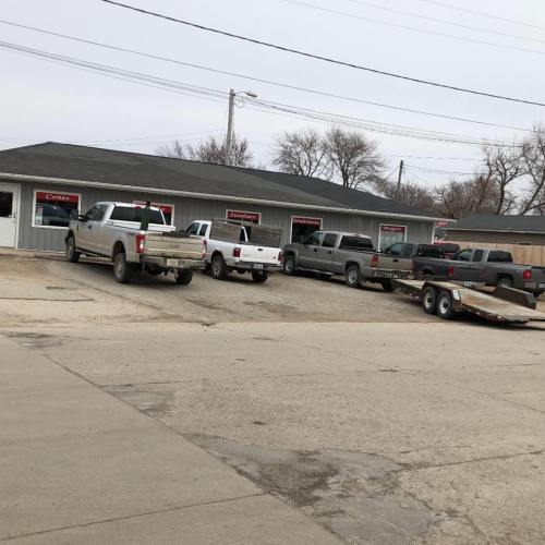 A row of pickup trucks parked outside a building with a gray exterior and signs, on a cloudy day.