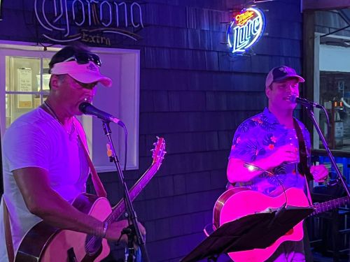 Two musicians perform with acoustic guitars under colorful lights at a lively venue.
