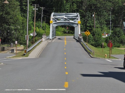 A view of a metal bridge over a road, surrounded by greenery and utility poles, with clear blue skies above.