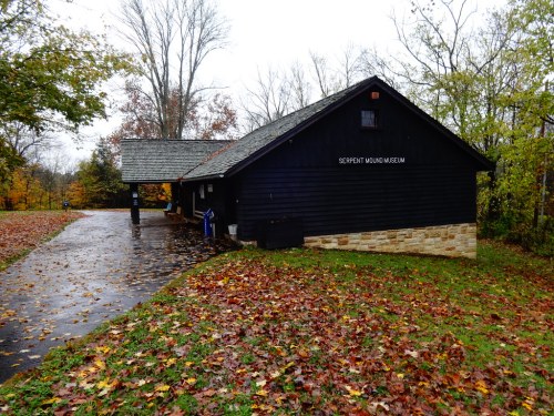 A dark wooden building with a stone base, labeled "Serpent Mound Museum," surrounded by autumn leaves and trees.