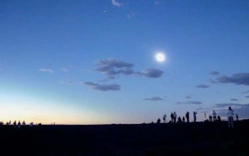 Silhouetted figures watch a solar eclipse against a twilight sky with clouds and a bright crescent moon.
