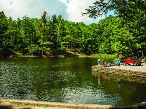 A serene lake surrounded by lush greenery, with people gathered by the water's edge and a red vehicle nearby.