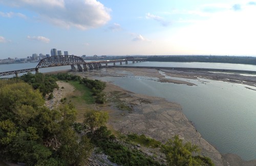 Aerial view of a river with a bridge, surrounded by dry land and greenery under a partly cloudy sky.
