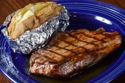 A grilled steak served on a blue plate alongside a baked potato wrapped in foil.