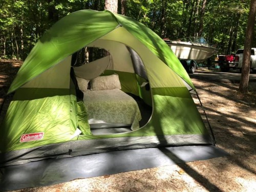 A green camping tent set up in a wooded area, with a bed inside and a boat parked nearby.