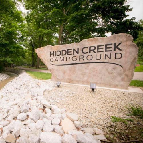 Sign for Hidden Creek Campground, surrounded by greenery and rocks, with a pathway visible in the background.