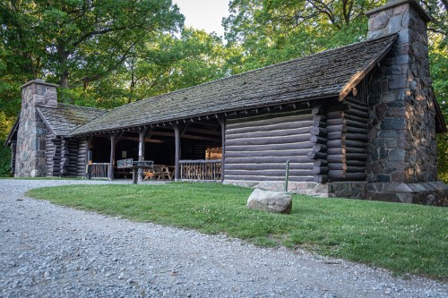 A rustic log cabin with stone accents, surrounded by greenery and a gravel path.