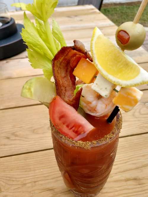 A vibrant Bloody Mary garnished with celery, bacon, shrimp, cheese, lemon, olive, and vegetables, served in a glass.