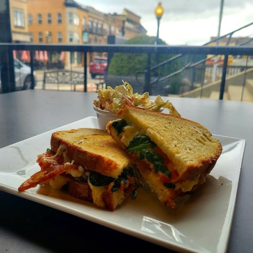 A grilled sandwich with bacon, spinach, and cheese, served with a side of coleslaw on a table with a street view.
