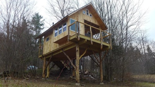 A wooden treehouse on stilts surrounded by trees, featuring a balcony and large windows.