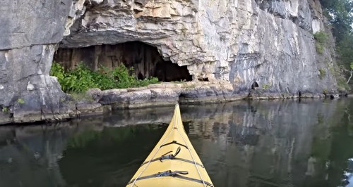 A yellow kayak approaches a rocky cliff with a cave entrance, surrounded by calm water and lush greenery.