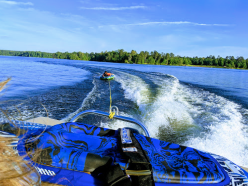 A view from a boat with a person being towed on a tube across a calm lake, surrounded by trees and blue sky.