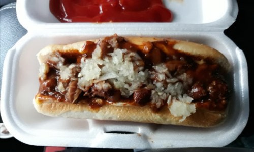 A hot dog topped with chopped onions and barbecue sauce, served in a takeout container with ketchup on the side.