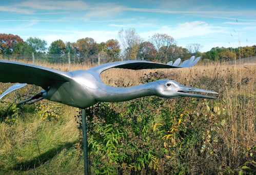 A metallic sculpture of a heron in a grassy field, with trees and blue sky in the background.