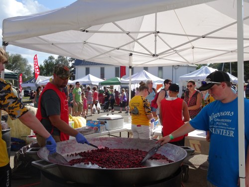 Volunteers prepare cranberries in a large pot at a festival, with tents and attendees in the background.