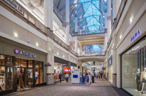 A spacious shopping mall interior with high ceilings, featuring stores like Athleta and Zara, and shoppers walking around.