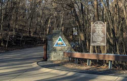 A stone entrance sign to a wooded area, featuring a triangular logo and a welcome message on a brown plaque.